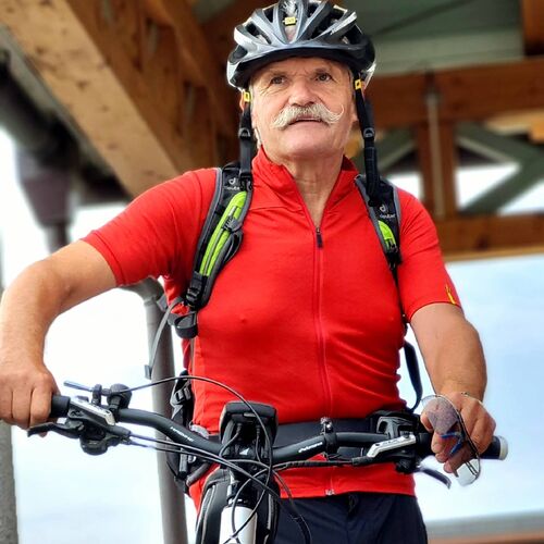 Älterer Mann mit Schnurrbart, Helm und rotem Trikot steht bereit mit E-Mountainbike vor dem Hotelstartpunkt.