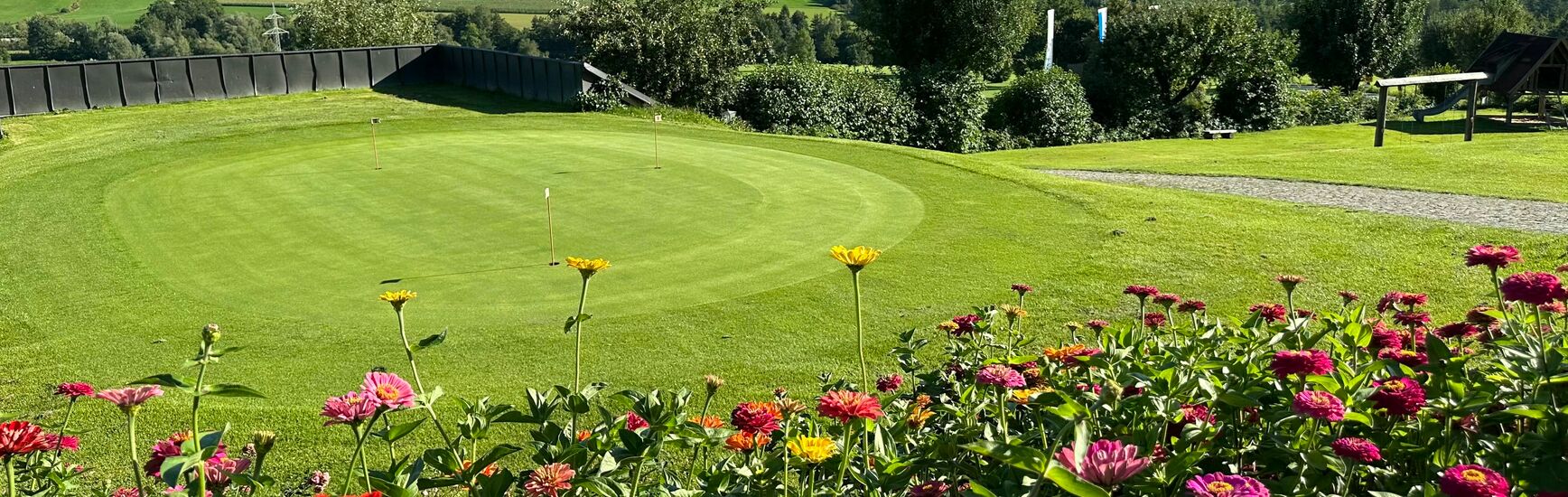Blick auf ein Putting-Green mit bunter Blumenrabatte im Vordergrund und weiten Ausblick auf Felder und Wälder.