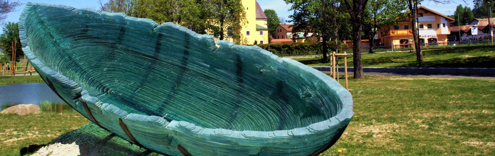 Grüne Glasskulptur in Form eines Bootes im Park der Gläsernen Gärten in Frauenau, im Hintergrund die gelbe Kirche mit Zwiebelturm.