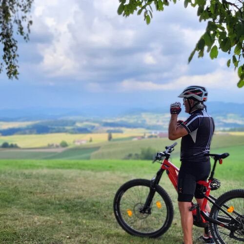 Radfahrer mit Helm betrachtet von einer Anhöhe aus die weite Landschaft des Bayerischen Waldes unter wolkigem Himmel.