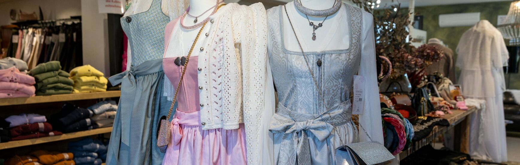 Drei Schaufensterpuppen tragen festliche Dirndl mit Spitzenschürzen und Accessoires im stilvoll eingerichteten Laden