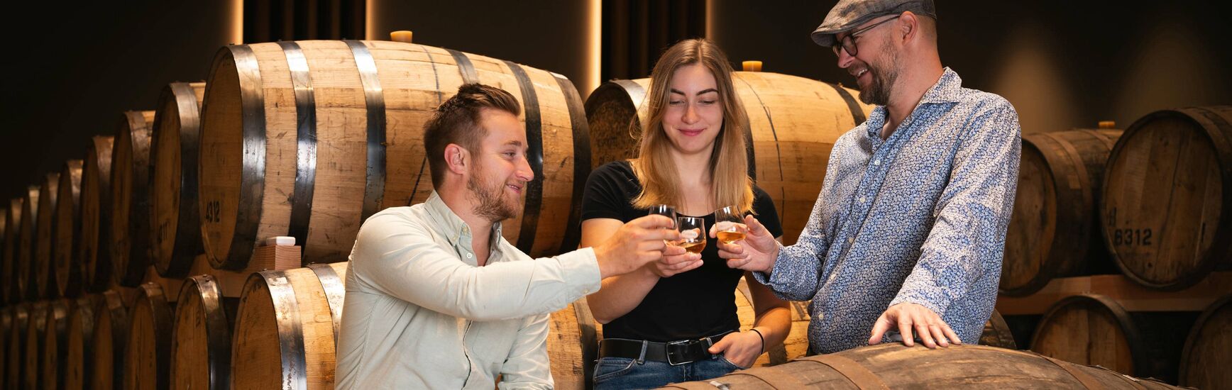 Drei Personen stoßen mit Whiskygläsern zwischen Holzfässern der Penninger Brennerei an.